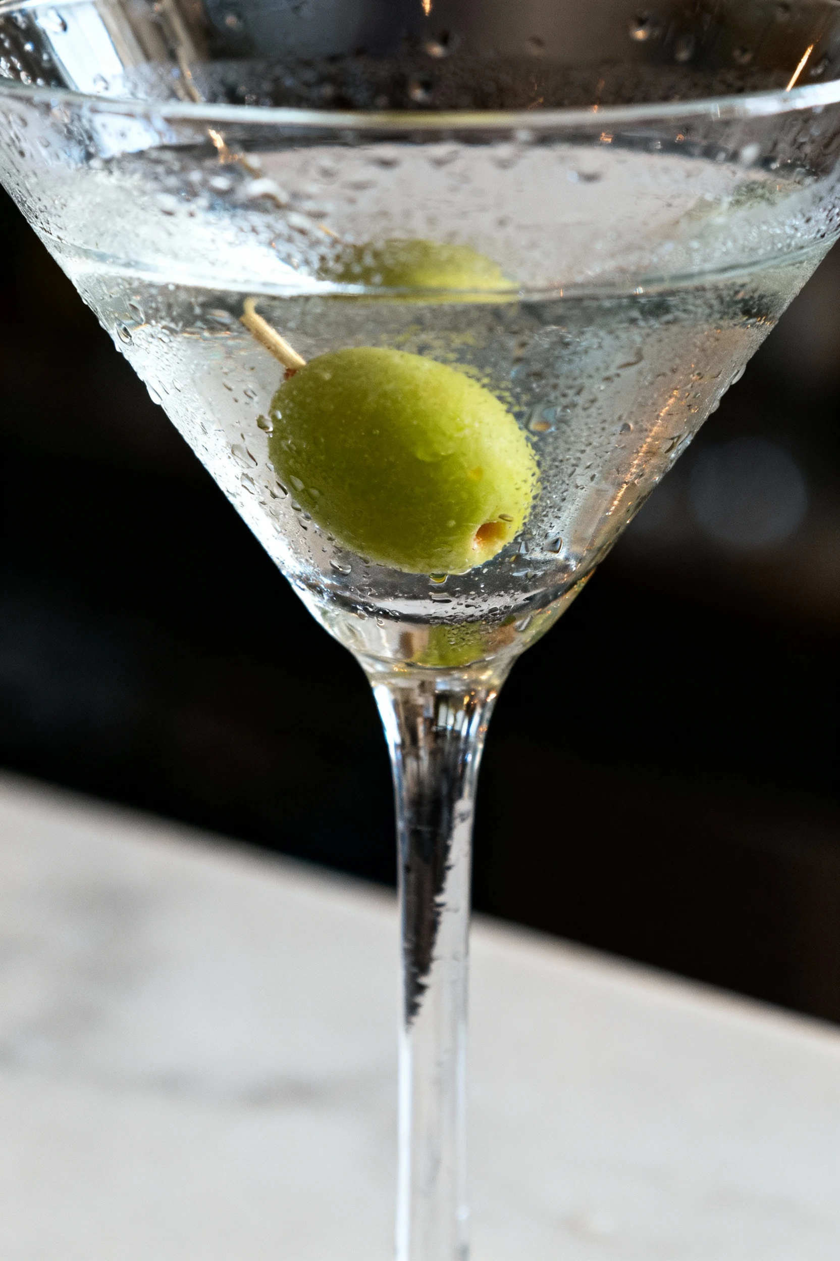 Close-up of a crystal-clear classic dry martini in a chilled stemmed glass, single green olive garnish, droplets of cond