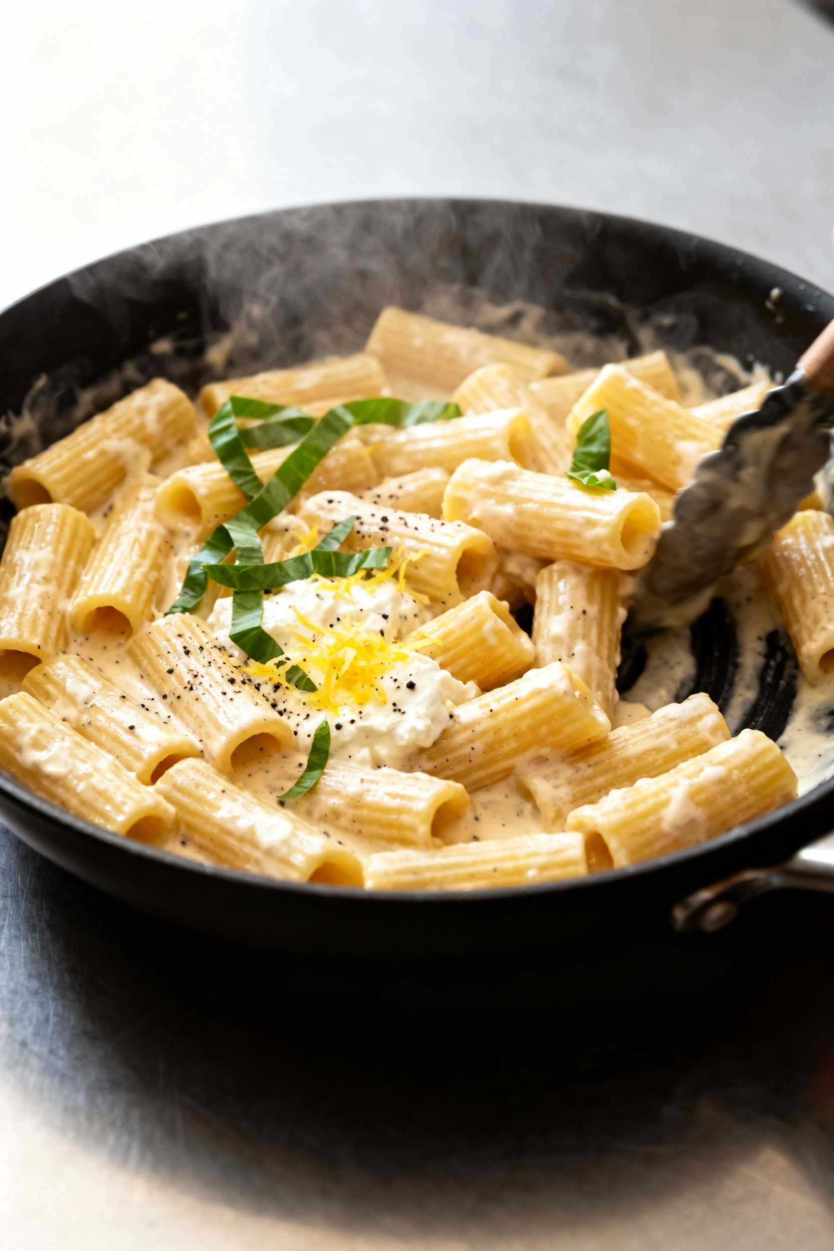 Lemon ricotta pasta being tossed in a skillet, glossy velvety sauce clinging to rigatoni with lemon zest, cracked black 