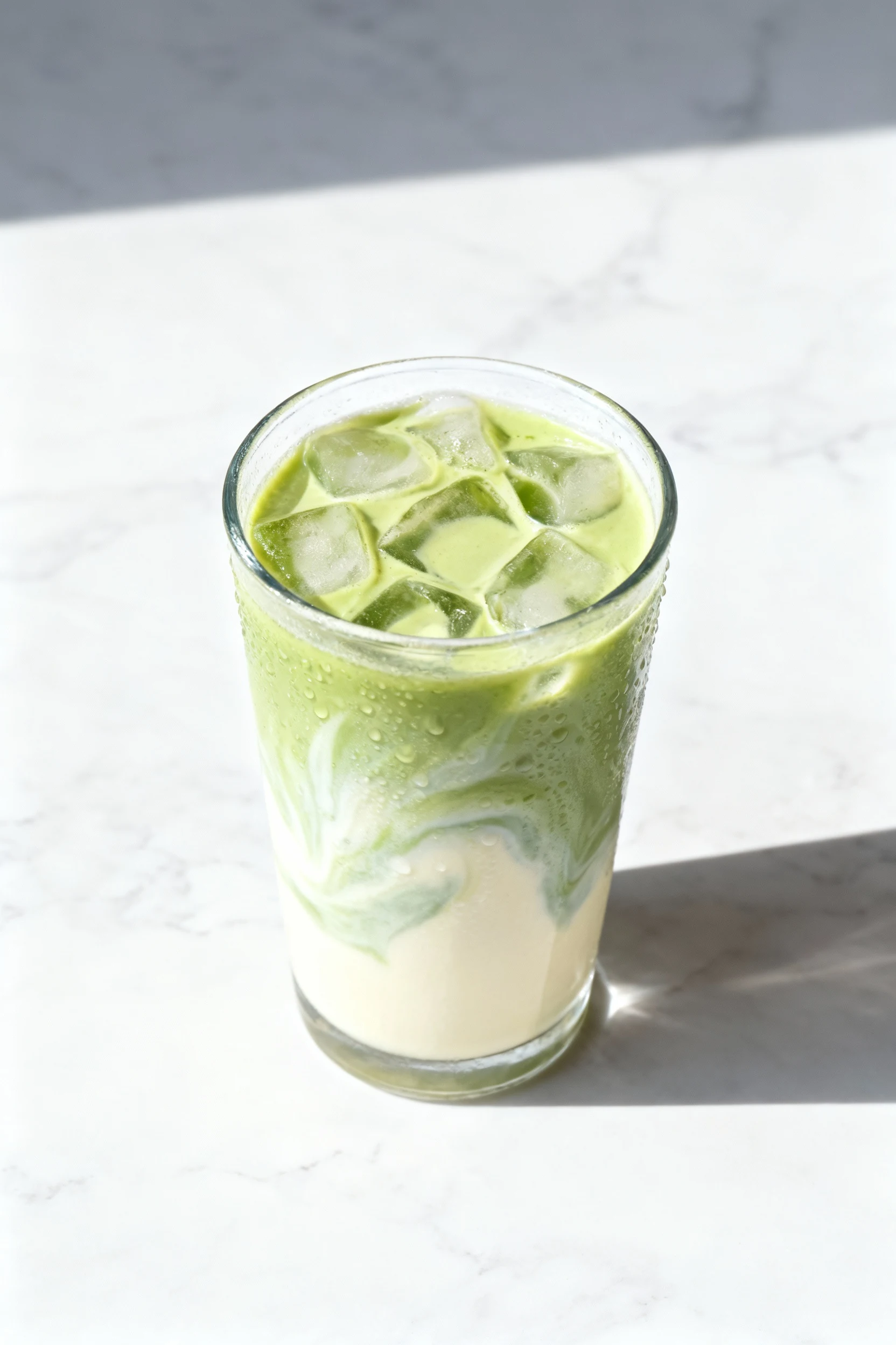 2. Overhead shot of iced matcha latte in a clear glass packed with ice, 1:4 concentrate-to-oat-milk swirl marbling pale 