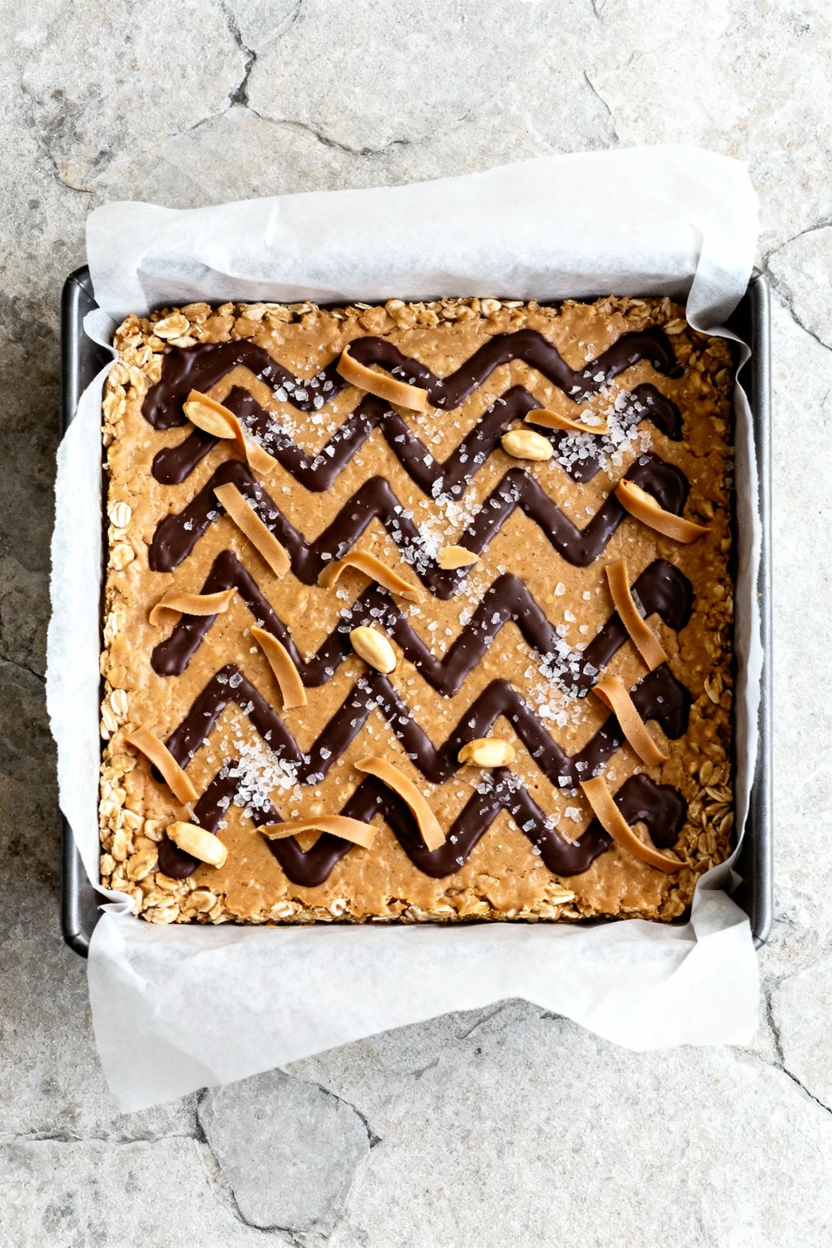 Overhead shot of an 8x8-inch parchment-lined pan with an evenly pressed peanut butter–oat layer, topped with zigzag dark