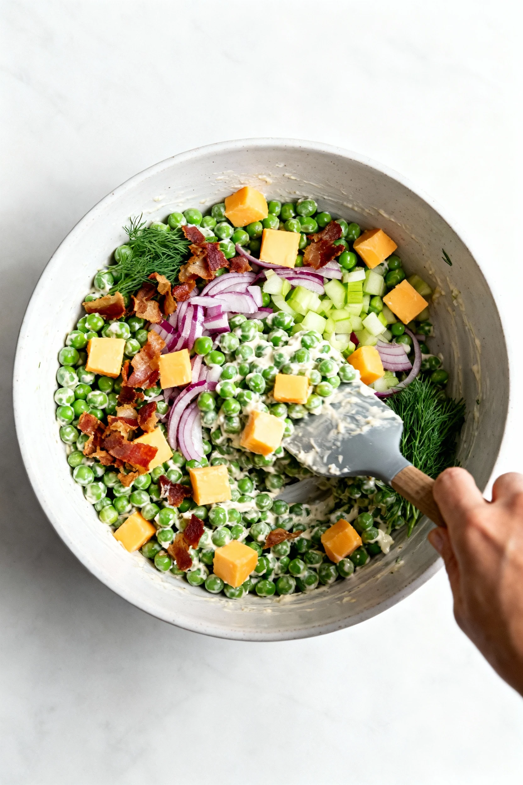 Process shot: pea salad being gently folded in a wide mixing bowl—intact peas with creamy dressing clinging, sharp chedd