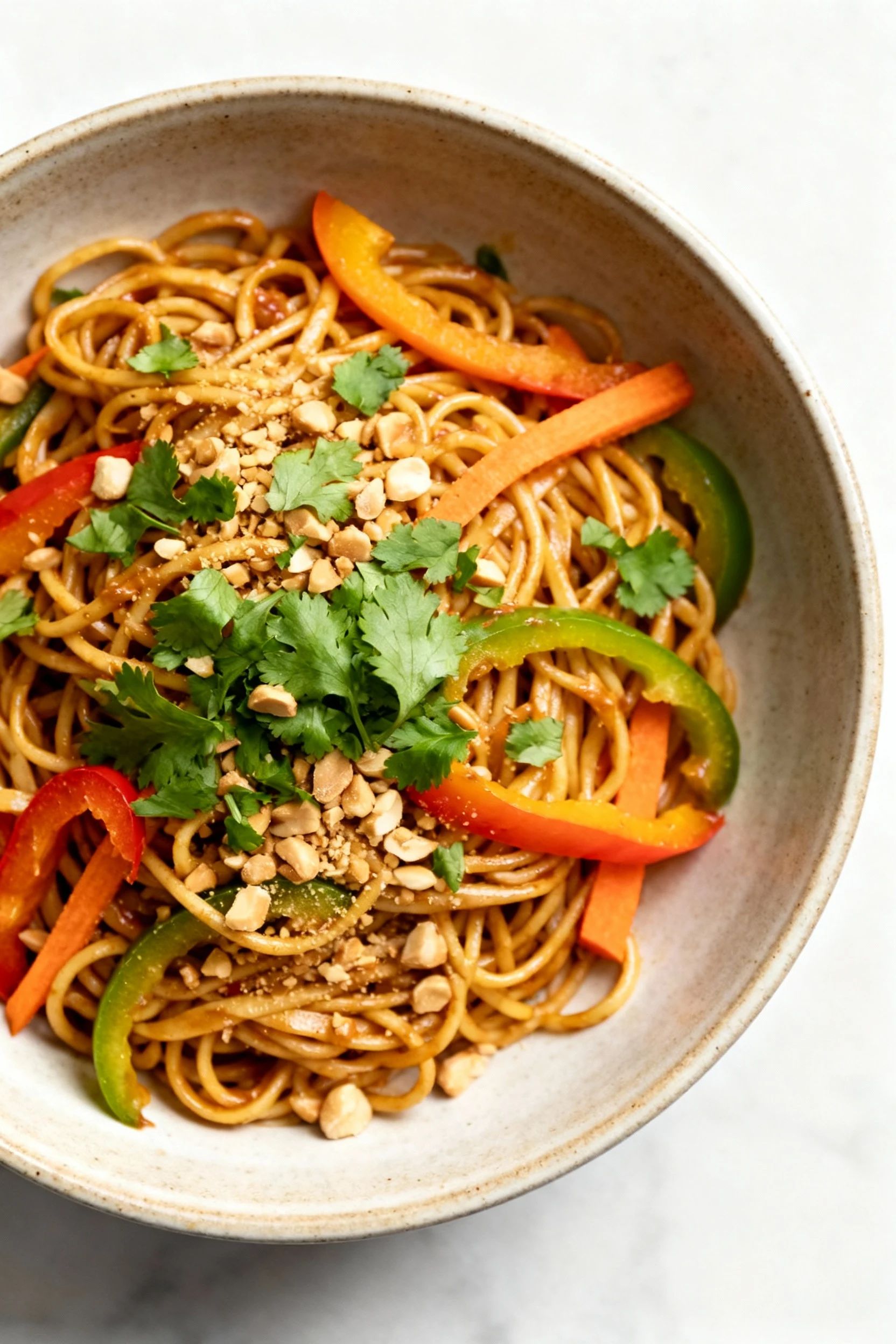 Overhead shot of vibrant Spicy Peanut Noodles topped with chopped cilantro and crushed peanuts, colorful bell pepper sli