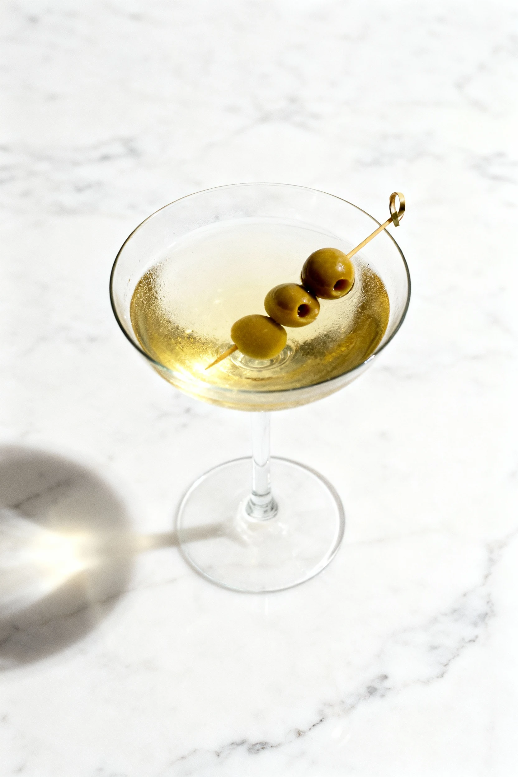 Overhead shot of a freshly strained dirty martini, pale golden hue from olive brine, three skewered olives resting elega