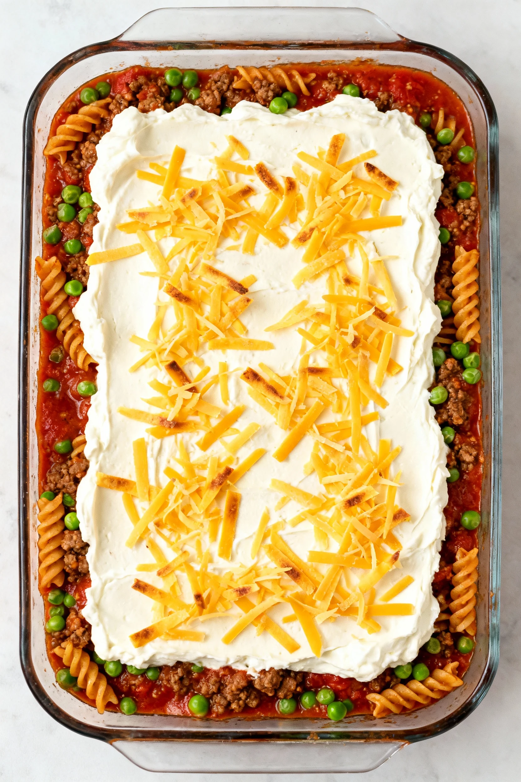 Overhead shot of assembled casserole in a 9x13 dish: beef-tomato sauce mixed with rotini and peas, creamy sour cream–cre