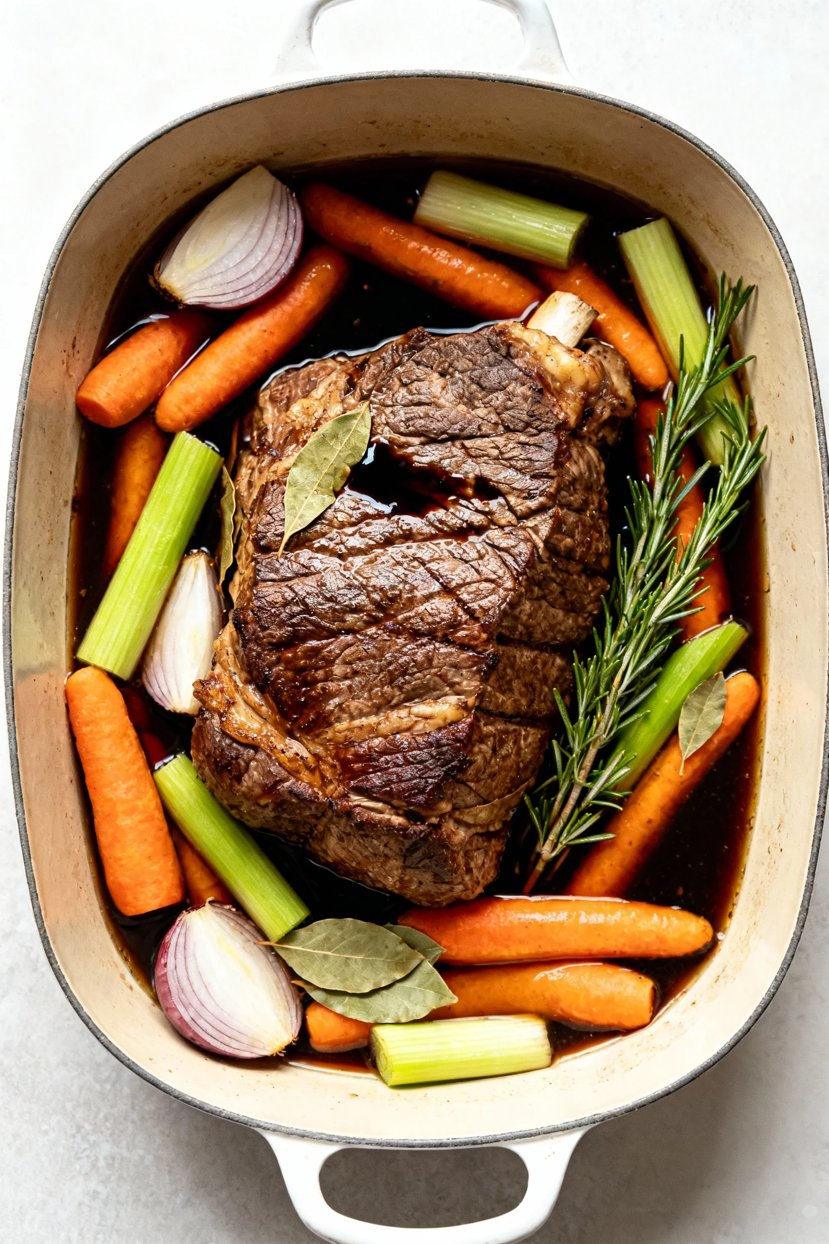3. Overhead mid-braise: roast nestled with carrots, onions, celery, bay leaves, rosemary; umami-rich broth (Worcestershi