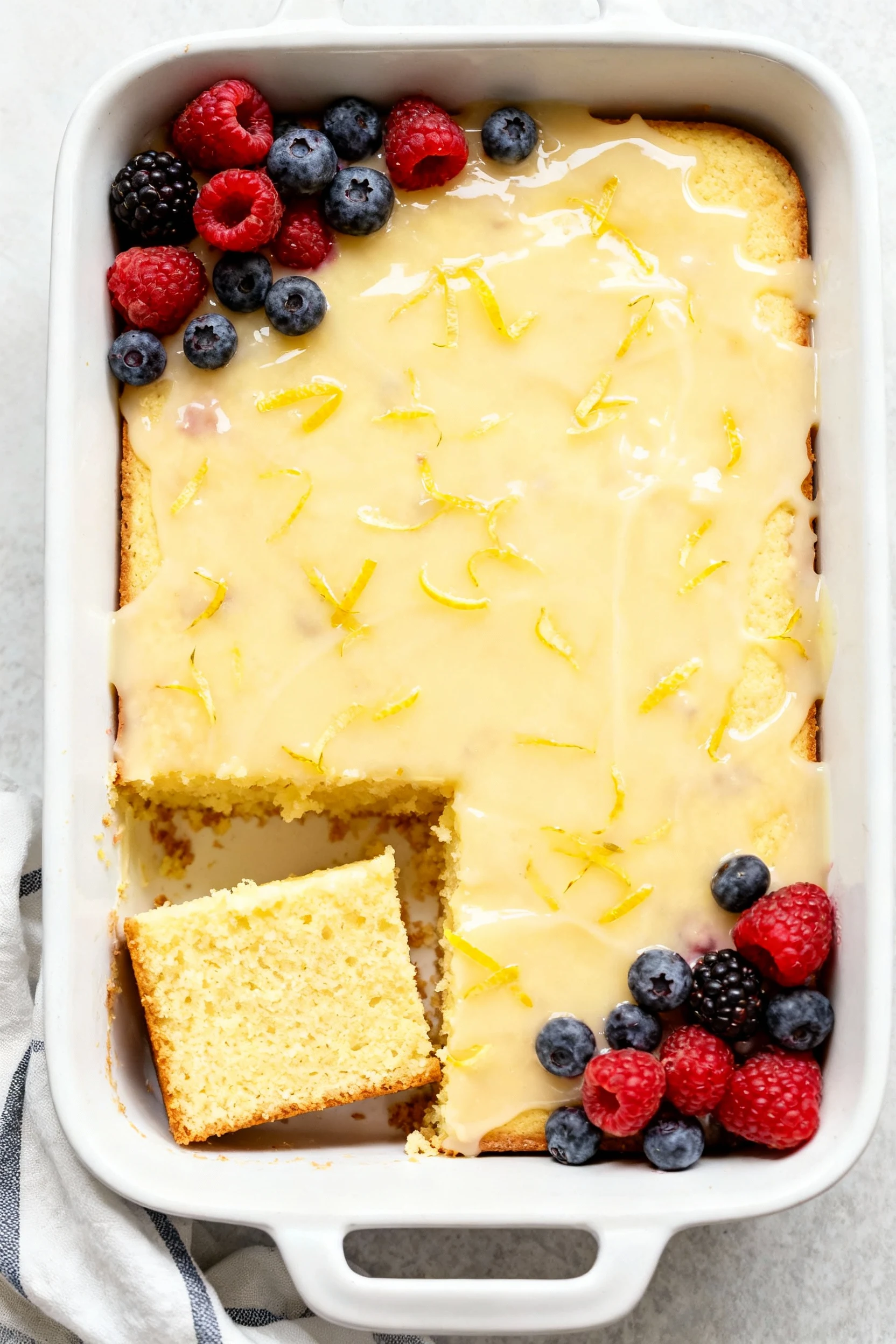 Overhead of Lemon Sunshine 13x9: pale-yellow cake under glossy lemon glaze with zest flecks and fresh berries; one squar