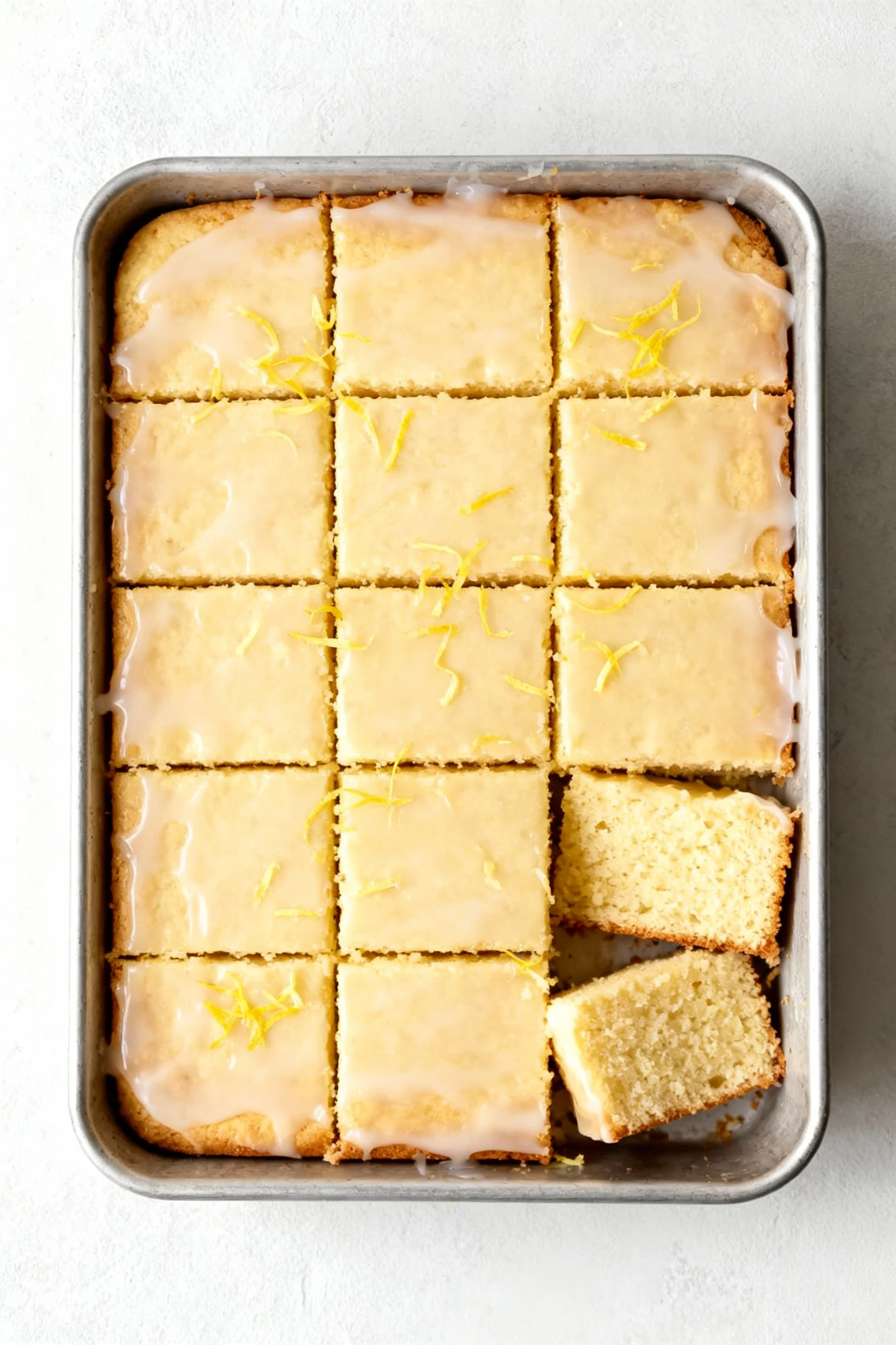 Overhead shot of 9×13 lemon yogurt sheet cake with a thin, translucent lemon glaze and fresh zest sprinkled on top; pale