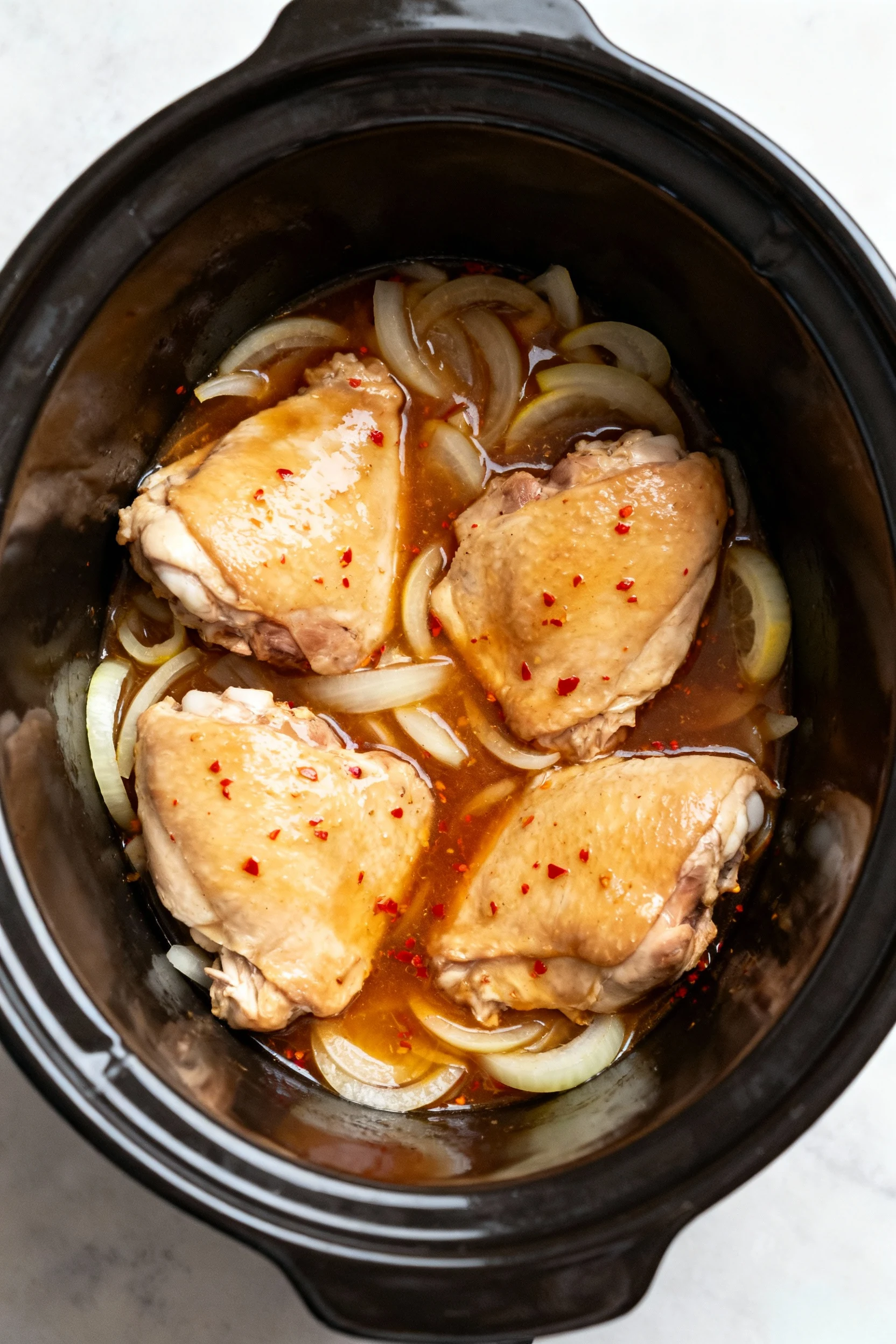 Overhead shot inside a slow cooker showing tender chicken thighs nestled on a bed of softened onions, bathed in amber ho