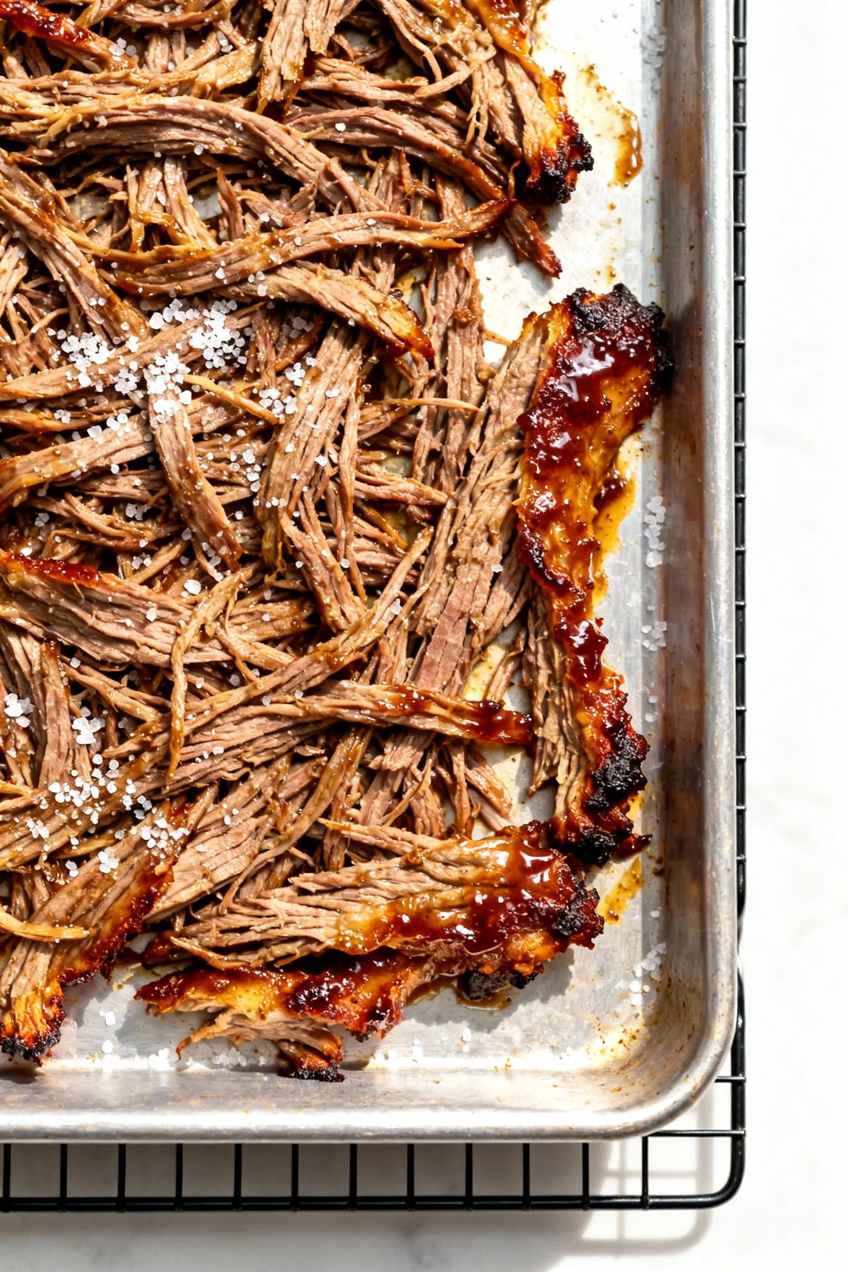 Cooking process detail: freshly broiled shredded beef spread thin on a sheet pan, edges caramelized and crackly, glossy 