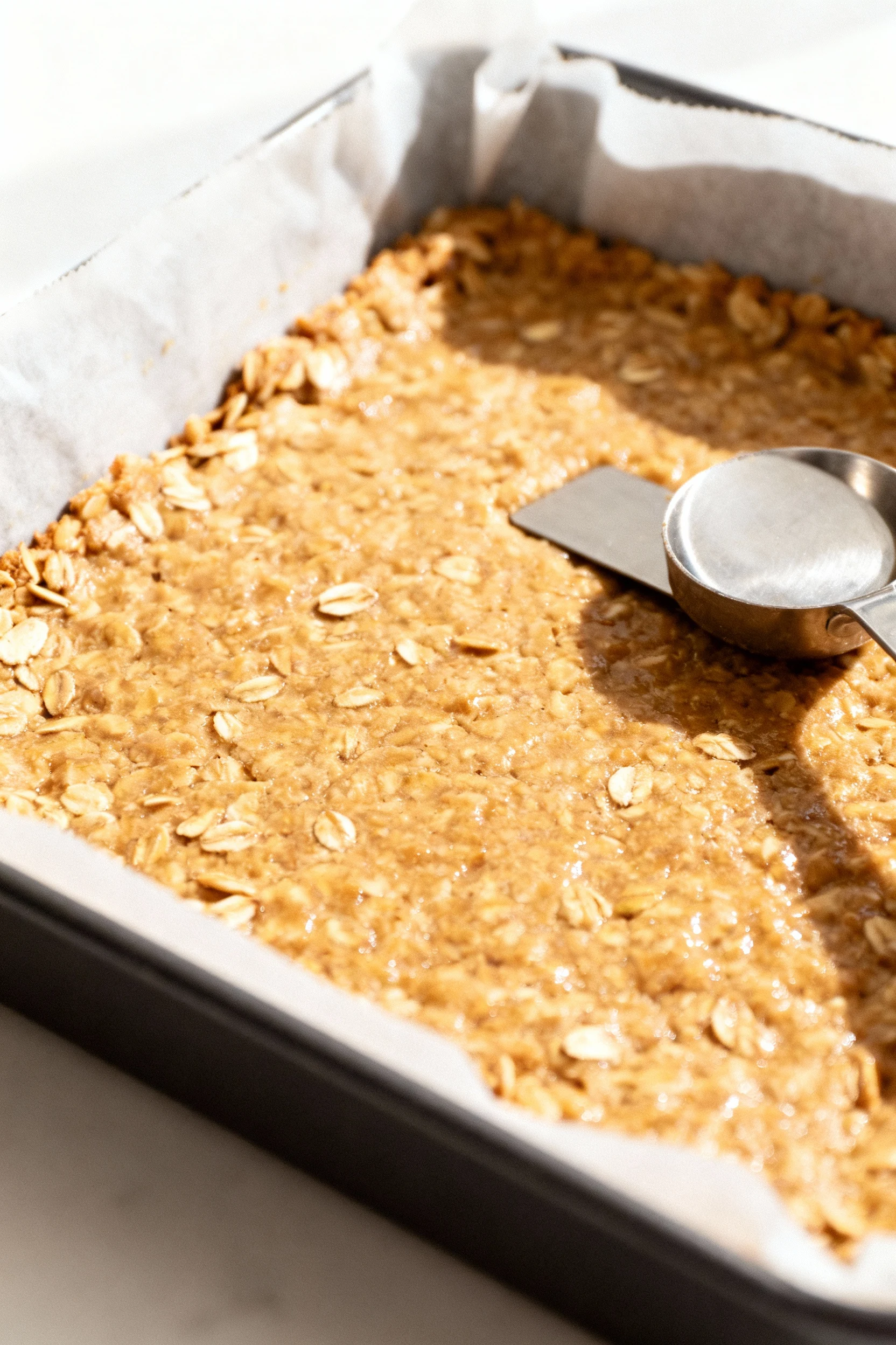 Process shot: compacting the warm oat mixture evenly in a lined square pan with the flat bottom of a metal measuring cup