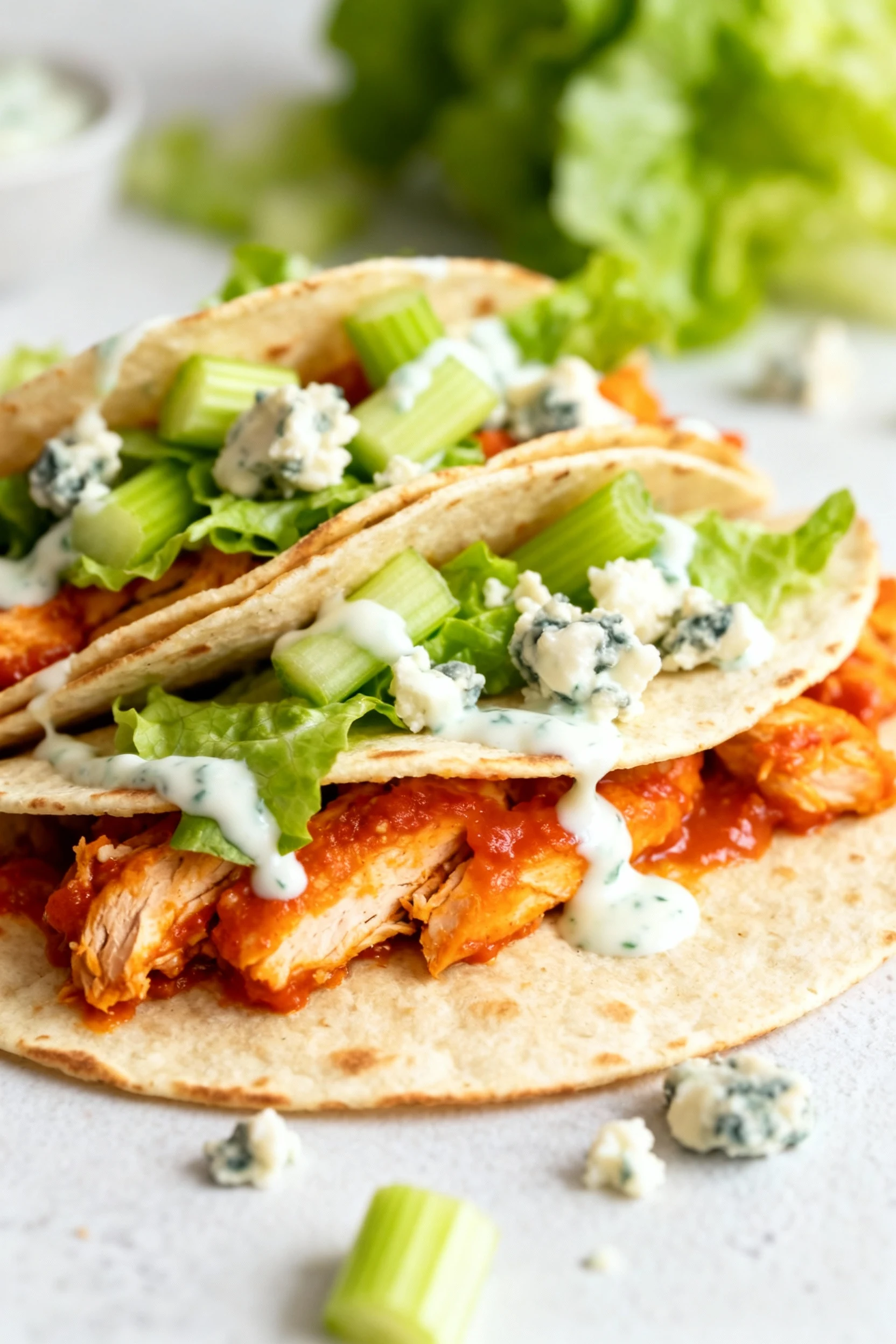 Close-up Buffalo chicken wraps: orange-red saucy chicken, crisp lettuce and celery, blue cheese crumbles, ranch drizzle;
