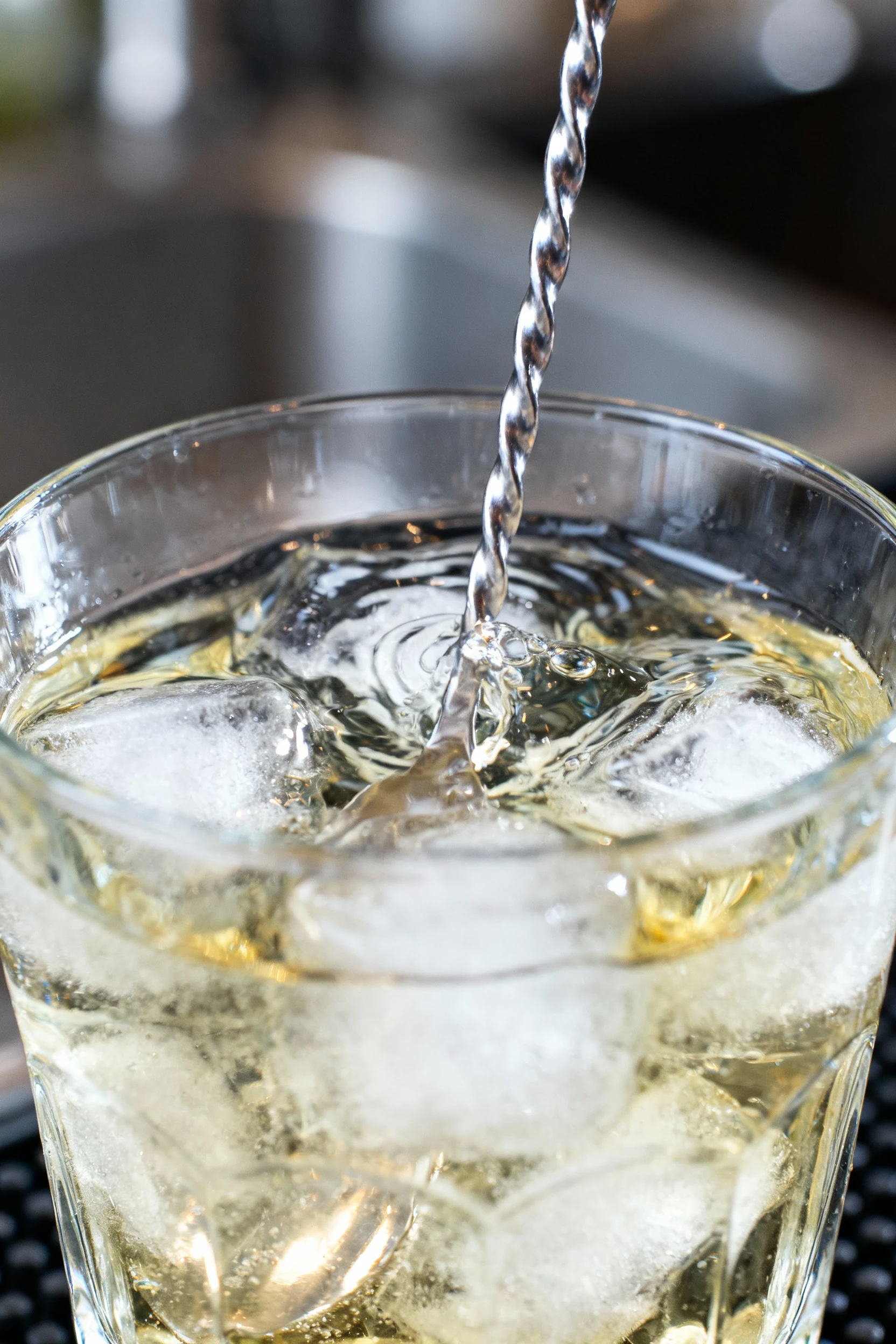 Professional kitchen scene showing gin and vermouth being stirred over ice in a mixing glass, the liquid swirling with g