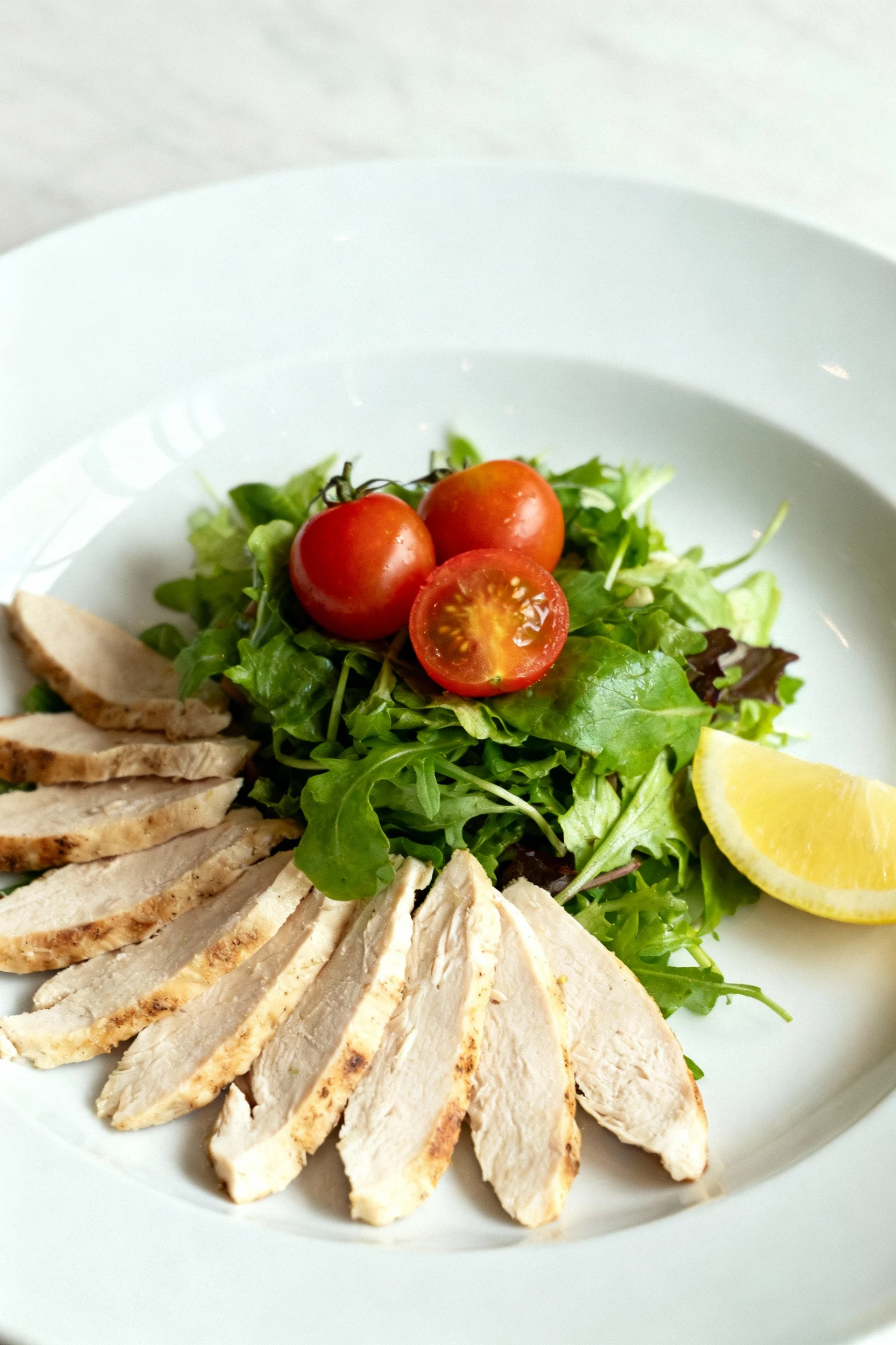 Elegant plated presentation of chicken salad on a wide white plate, chicken fanned across greens, warm tomatoes placed o