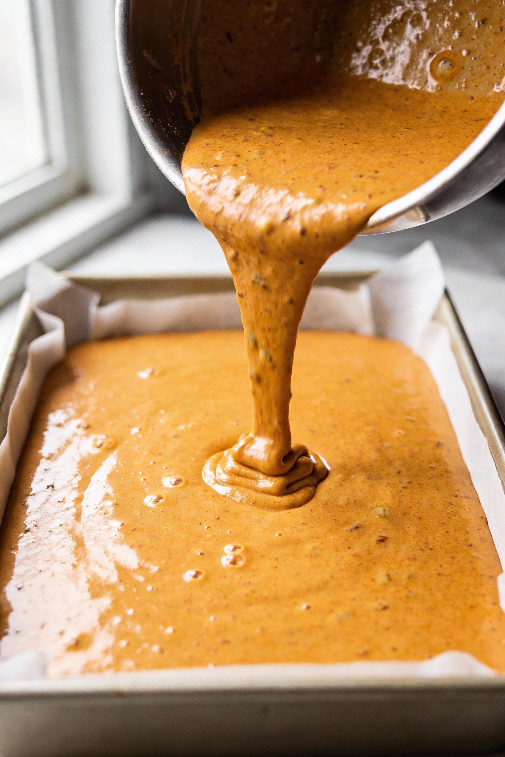 Pumpkin cake batter being poured into a parchment-lined light-colored metal 9x13 pan, silky orange batter with cinnamon-