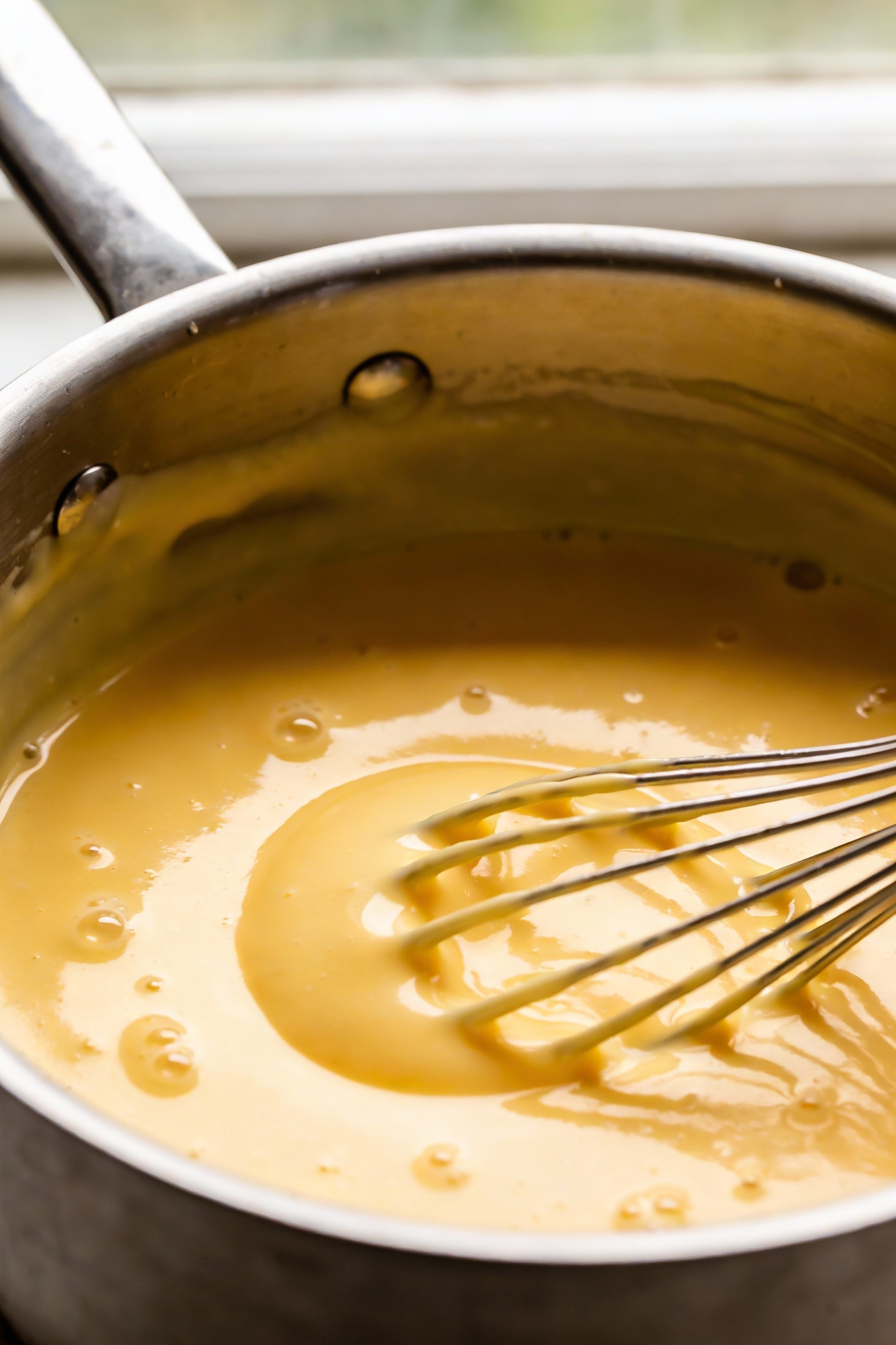 Close-up detail of vanilla custard thickening in a stainless saucepan, glossy pudding-like texture with visible whisk tr