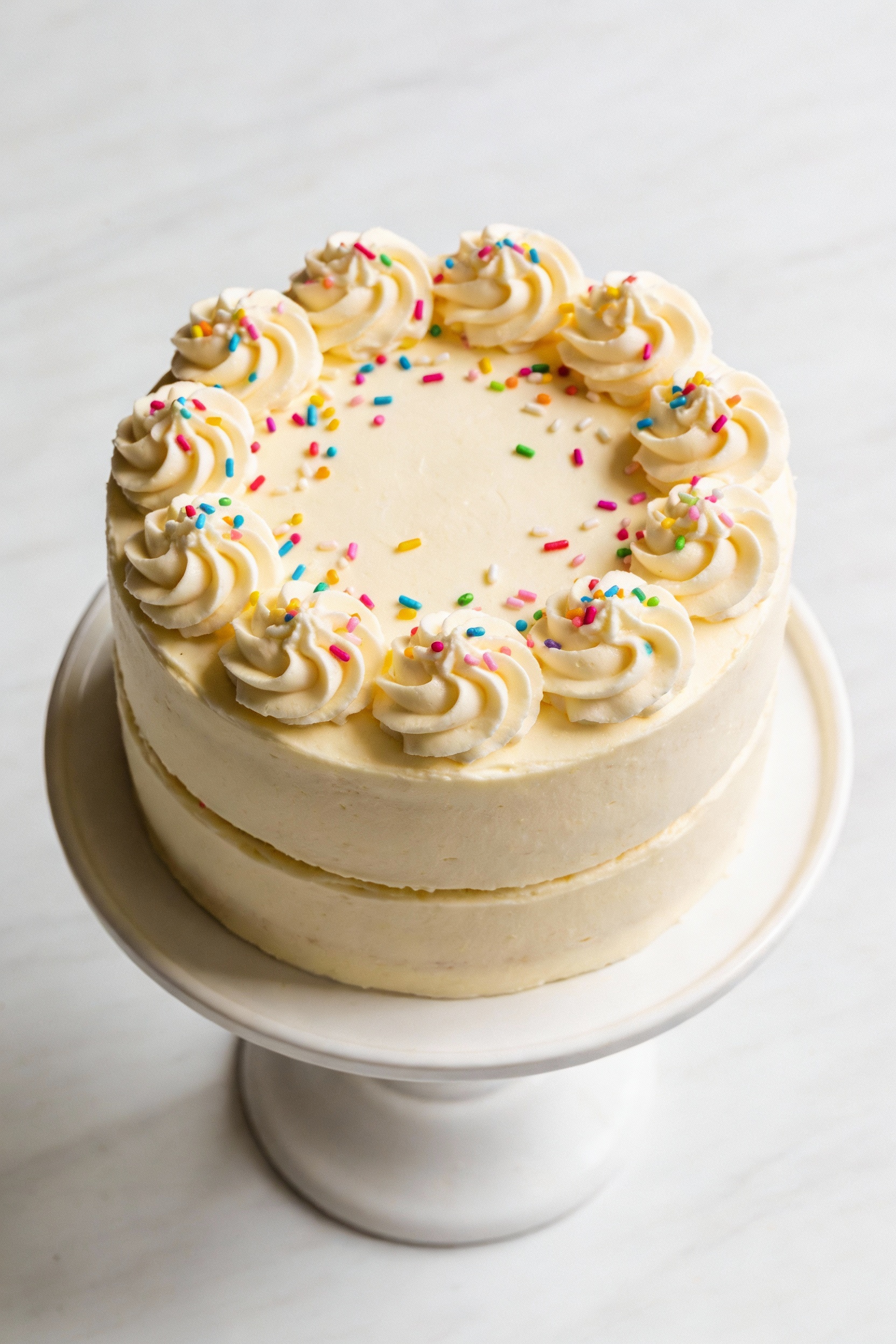 Overhead shot of a fully frosted two-layer vanilla celebration cake on a white cake stand, smooth sides with clean edges