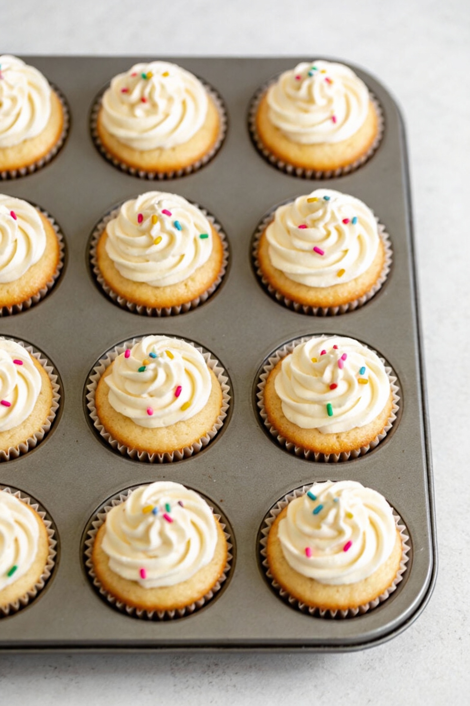 2. Overhead shot of a 12-cup muffin tin filled with baked vanilla cupcakes (doctored cake mix), evenly domed tops with g