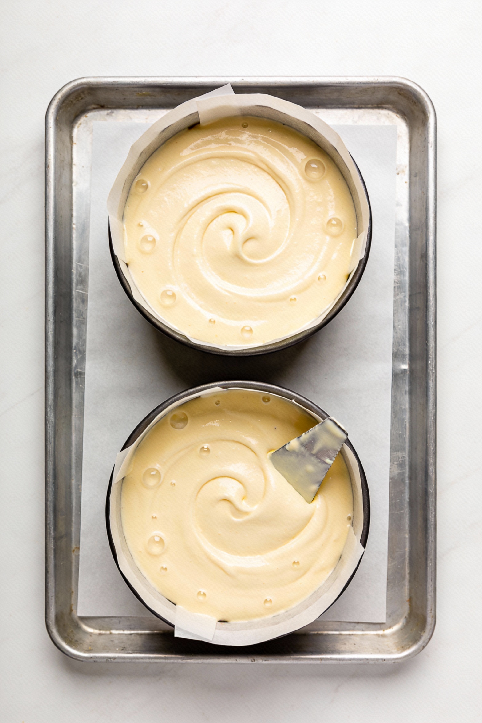 Cooking process: reverse-creamed vanilla cake batter being smoothed in two parchment-lined 8-inch round pans, silky pale