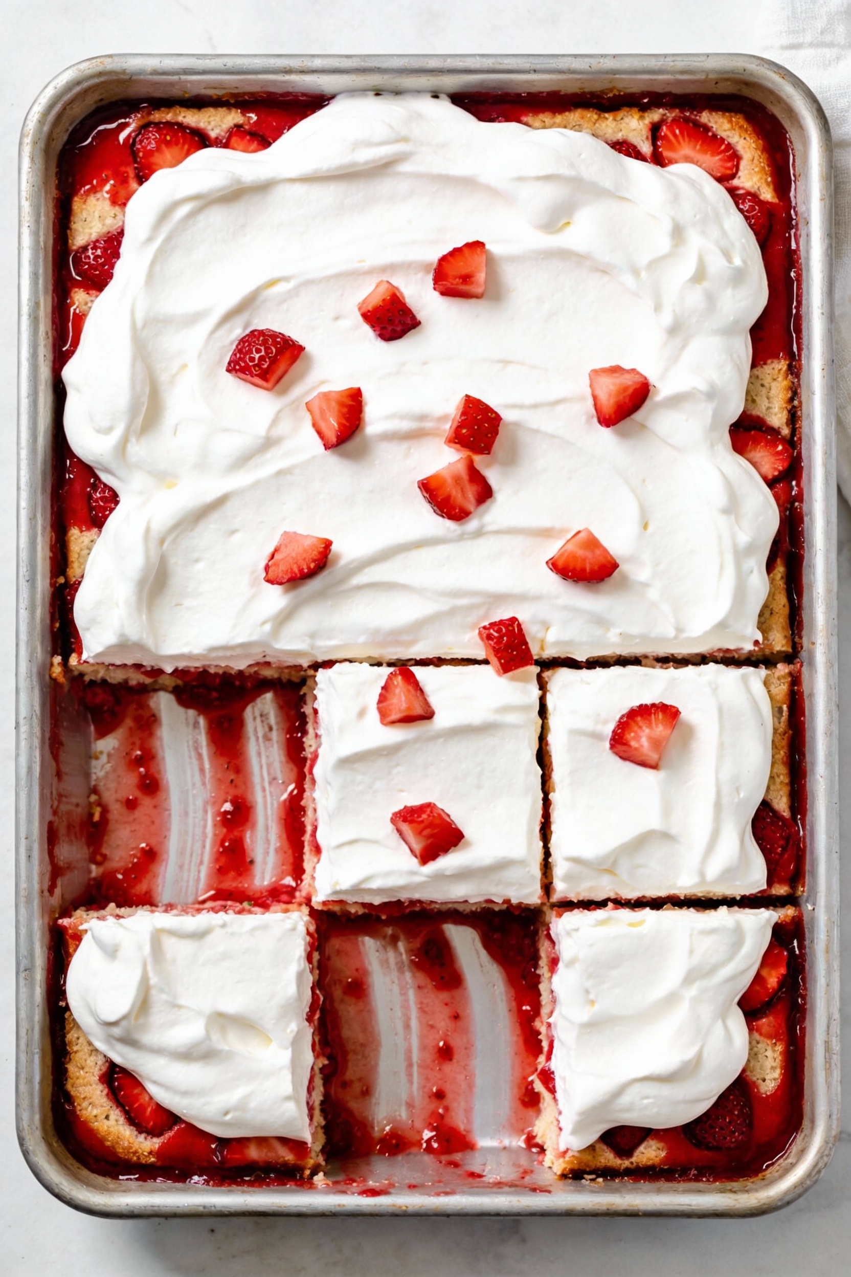 Overhead of strawberries & cream poke cake in a 9x13 pan, clean slices revealing strawberry puree ribbons beneath billow