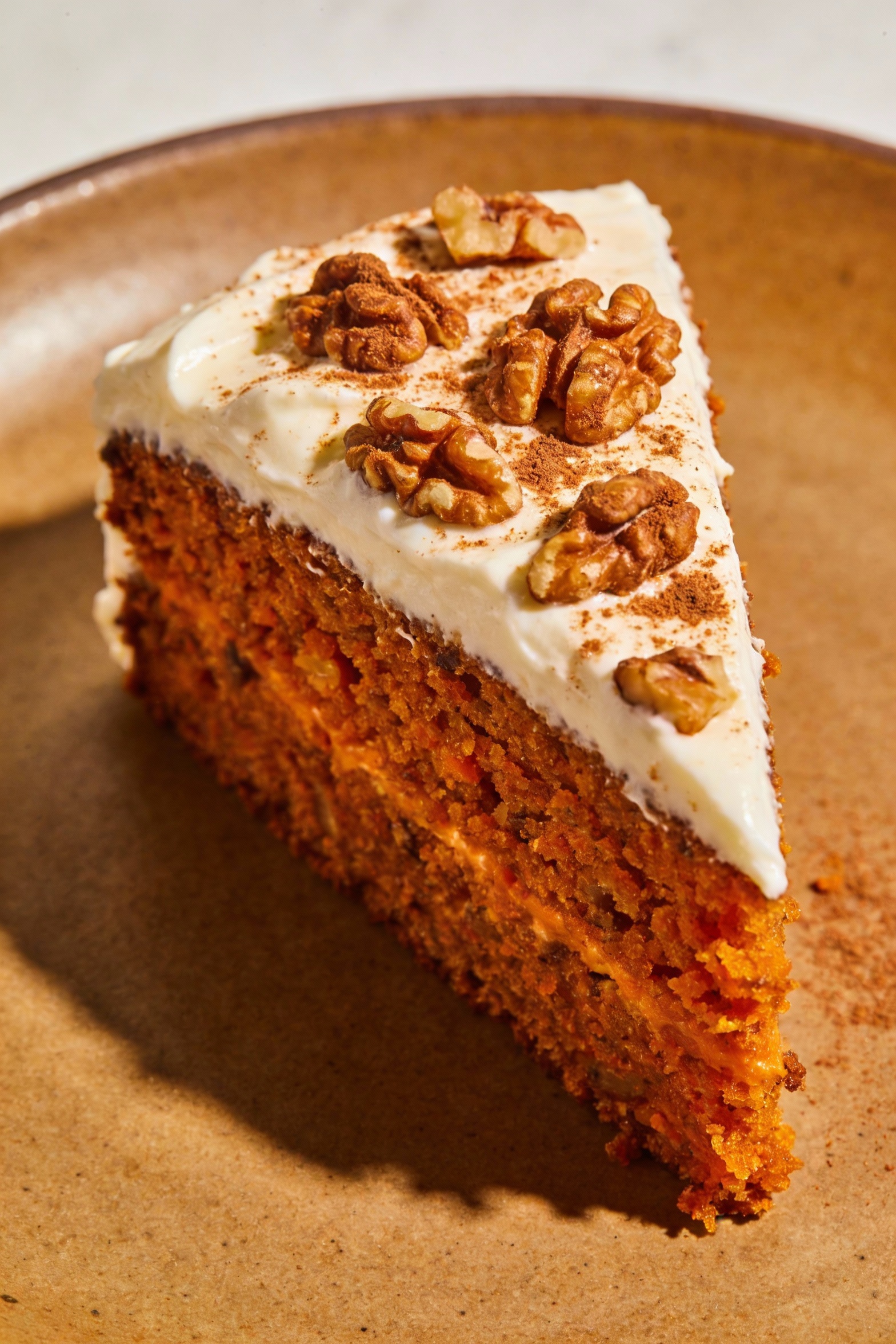 Beautifully plated carrot spice cake slice with thick cream cheese frosting, topped with toasted walnut pieces and a dus